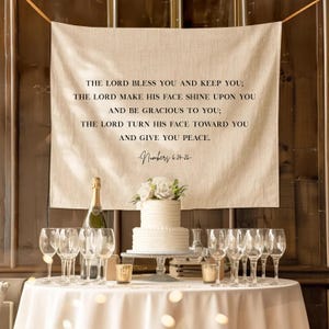 May include: A wedding reception scene featuring a white cake with floral decorations, champagne, and wine glasses. A banner with a religious quote hangs above the table. The table is set for a celebration.