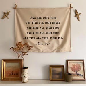 May include: A beige fabric wall hanging with the text "LOVE THE LORD YOUR GOD WITH ALL YOUR HEART AND WITH ALL YOUR SOUL AND WITH ALL YOUR MIND AND WITH ALL YOUR STRENGTH. Mark 12:30." Two gold bird wall decorations flank the hanging. A shelf below holds framed art and a vase.