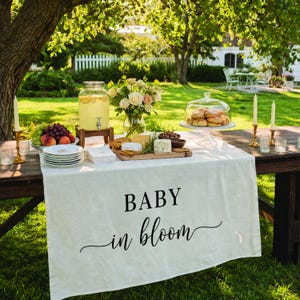 May include: Outdoor table setting with a white tablecloth featuring the text "BABY in bloom." The table is laden with food and drinks, including a glass beverage dispenser, cheese, crackers, fruit, and a cake under a glass dome. The scene is set in a garden.
