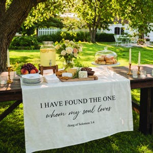 May include: A wooden table set for an outdoor gathering, featuring a white tablecloth with the text "I HAVE FOUND THE ONE whom my soul loves -Song of Solomon 3:4". The table holds food, drinks, and flowers, including a glass beverage dispenser, plates, and a cake stand.