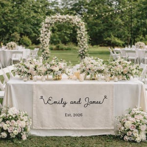 May include: A wedding reception table is set with a white tablecloth and floral arrangements. A runner displays the names "Emily and James" and the date "Est. 2026". The scene includes an archway decorated with flowers and additional tables.