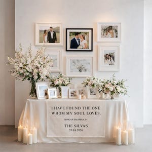 May include: A wedding reception display featuring framed photos, floral arrangements, and candles. A table is draped with a cream-colored cloth, adorned with a banner that reads "I HAVE FOUND THE ONE WHOM MY SOUL LOVES." The date 25.04.2026 is also displayed.