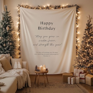 May include: A cream-colored tapestry with the words "Happy Birthday" and a quote. The tapestry is surrounded by fairy lights and greenery. Two decorated Christmas trees flank the tapestry. A small table holds candles and pinecones, with wrapped gifts nearby.