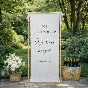 May include: A cream-colored banner with the text "FOR THIS CHILD We have prayed -1 Samuel 1:27-" is displayed on a wooden frame. A basket of white flowers and a basket of champagne bottles are on either side of the banner. The background is a garden setting.