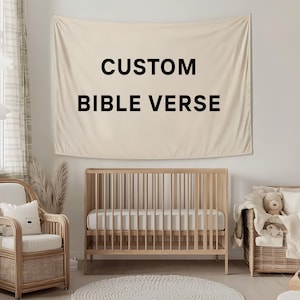 May include: A beige wall tapestry with the words "CUSTOM BIBLE VERSE" in black lettering hangs above a wooden crib. The room also features a wicker chair, a round rug, and a wicker storage basket.