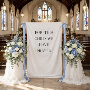May include: A white banner with the text "FOR THIS CHILD WE HAVE PRAYED" is displayed in a church. The banner is supported by light blue ribbons and flanked by floral arrangements of white roses, blue hydrangeas, and greenery.
