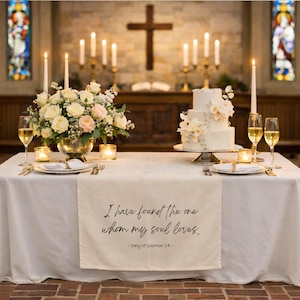May include: A wedding reception table is set with a white tablecloth, floral arrangements, candles, and a tiered cake. A table runner displays the text "I have found the one whom my soul loves."
