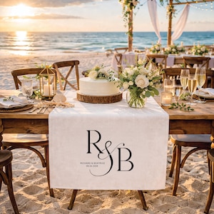 May include: A beach wedding reception setup with a white cake on a wooden base, floral arrangements, and champagne glasses. A white table runner displays the initials "R & B" and the date "09.04.2026". The scene is set against the ocean.