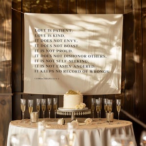 May include: A wedding reception scene with a white cake adorned with gold and white flowers on a decorative stand. A banner with the text "LOVE IS PATIENT" hangs above the table. Champagne glasses and candles are arranged around the table.