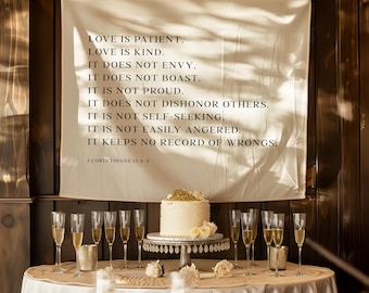 Puede incluir: Escena de recepción de boda con una tarta blanca adornada con flores doradas y blancas sobre un soporte decorativo. Una pancarta con el texto "LOVE IS PATIENT" cuelga sobre la mesa. Copas de champán y velas están dispuestas alrededor de la mesa.