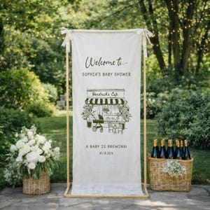 May include: A cream-colored banner with the text "Welcome to SOPHIA'S BABY SHOWER" and a drawing of a cafe with a stroller. The banner is held by a wooden frame and tied with white ribbons. The text "A BABY IS BREWING!" and the date 05.10.2026 are also visible.