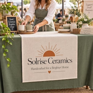May include: A beige table runner featuring a burnt orange sun graphic and the text Solrise Ceramics Handcrafted for a Brighter Home with a small heart icon. The runner is draped over a green tablecloth, with ceramic mugs and decor nearby.