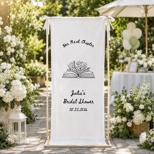 May include: A tall, rectangular white fabric banner suspended from a wooden frame. The banner features black text reading Her Next Chapter, a line drawing of an open book with flowers, and the text Julia's Bridal Shower with the date 08.22.2026.