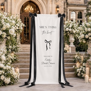 May include: A vertical white fabric banner featuring black text that reads "SHE'S TYING the knot" above a black bow graphic and "WELCOME TO Emily's Bridal Shower 08.22.2026". The banner is accented with long black ribbons tied into bows at the top corners.