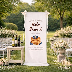 May include: A white banner with the words "Welcome to Samantha's Baby Brunch" and "August 18 2026" in brown script. The banner has an illustration of pancakes with blueberries and syrup. The banner is hanging outdoors.