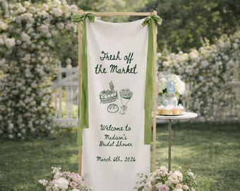 May include: A white banner with the text "Fresh off the Market" and illustrations of produce, flowers, and bread. The banner is part of a wooden frame, adorned with green ribbons, and welcomes guests to Madison's Bridal Shower on March 6th, 2026.