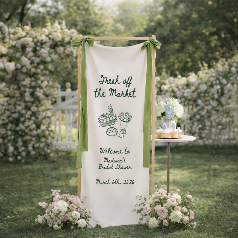 May include: A white banner with the text "Fresh off the Market" and illustrations of produce, flowers, and bread. The banner is part of a wooden frame, adorned with green ribbons, and welcomes guests to Madison's Bridal Shower on March 6th, 2026.