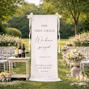 May include: A white banner with the text "FOR THIS CHILD We have prayed -1 Samuel 1:27-" is displayed at an outdoor baby shower. The banner is decorated with ribbons and is next to a table with wine bottles and glasses.
