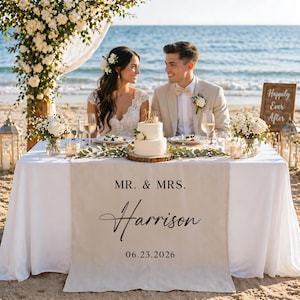 May include: A beach wedding reception setup with a white tablecloth, floral arrangements, and a two-tiered cake. A linen table runner reads "MR. & MRS. Harrison 06.23.2026". A wooden sign says "Happily Ever After". A couple is seated at the table.