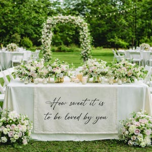 May include: A beautifully decorated outdoor wedding reception. A table is set with a white tablecloth, floral arrangements, and candles. A banner on the table reads, "How sweet it is to be loved by you."