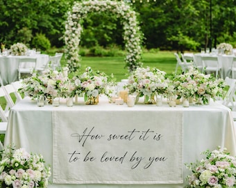 May include: A beautifully decorated outdoor wedding reception. A table is set with a white tablecloth, floral arrangements, and candles. A banner on the table reads, "How sweet it is to be loved by you."