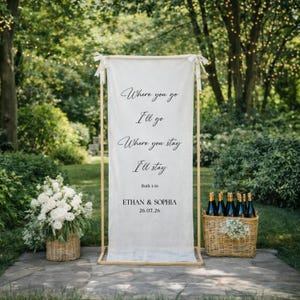 May include: A wedding sign with the text "Where you go I'll go, Where you stay I'll stay, Ruth 1:16, Ethan & Sophia, 26.07.26". The sign is on a wooden frame. A basket of flowers and a basket of champagne bottles are on either side.