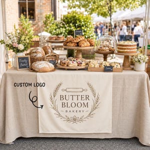 May include: A beige linen table runner featuring a custom logo with the text BUTTER BLOOM BAKERY, a rolling pin, and wheat stalks. The table is filled with assorted baked goods including loaves of bread, croissants, scones, and a layered cake.