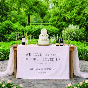 May include: A wedding reception table set outdoors with a three-tiered cake, wine bottles, champagne, and candles. A white tablecloth displays the text "WE LOVE BECAUSE HE FIRST LOVED US -1 David 5:20- GLORIA & JORDAN 07.26.2026."