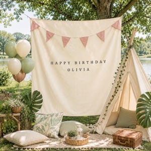 May include: A cream-colored fabric backdrop featuring the black text HAPPY BIRTHDAY OLIVIA and a pink triangular bunting banner. The scene includes a canvas play tent, assorted sage green and cream balloons, decorative pillows, and a wicker basket.