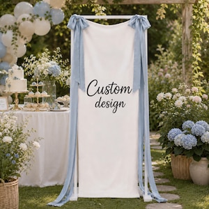 May include: A rectangular white fabric sign held by a white frame, featuring the black text Custom design. The sign is decorated with two long, light blue satin ribbons tied in bows at the top corners, draping down the sides of the frame.