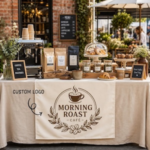 May include: A beige table runner featuring a brown printed logo that reads Morning Roast Café with a coffee cup icon and leaf wreath. The runner is displayed on a table with a coffee machine, coffee bags, pastries, and signs reading Coffee Tea Pastries.