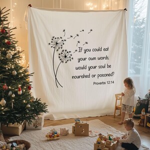 May include: A large, off-white fabric wall hanging with a black dandelion design and the text "If you could eat your own words, would your soul be nourished or poisoned? Proverbs 12:14." A Christmas tree and children playing are in the background.