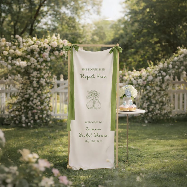 Puede incluir: Una pancarta blanca con cintas verdes y un marco de madera, expuesta al aire libre. La pancarta dice "She found her Perfect Pear" con una ilustraci&oacute;n de pera y "Welcome to Emma's Bridal Shower May 25th, 2026."