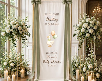 May include: A vertical white fabric banner on a gold metal frame, featuring the text A LITTLE SWEET Duckling IS ON THE WAY, a duck illustration with balloons, and WELCOME TO Olivia's Baby Shower 12.07.2026, accented with sage green ribbons.