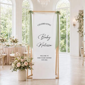 May include: A tall white fabric banner suspended from a wooden frame with sage green ribbons tied at the top corners. The banner features black text reading CELEBRATING Baby Melissa, WELCOME TO EMILY'S BABY SHOWER, and the date 19.07.2026.