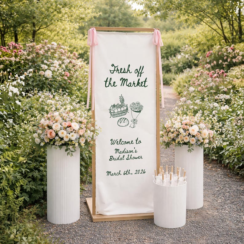 May include: An outdoor scene with a wooden framed sign that reads "Fresh off the Market" and "Welcome to Madison's Bridal Shower March 6th, 2026". The sign is flanked by floral arrangements on white pedestals. Pink ribbons adorn the top.