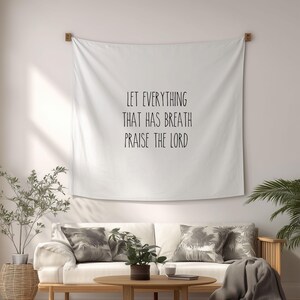 May include: White wall tapestry with the text "LET EVERYTHING THAT HAS BREATH PRAISE THE LORD" in black. The tapestry is hanging above a white sofa with decorative pillows. A wooden coffee table and plants are in front of the sofa.