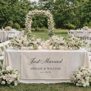 May include: A wedding reception table is decorated with floral arrangements and candles. A linen runner displays the text "Just Married" with the names Abigail & William and the year 2026. The scene is set outdoors with an archway of flowers.