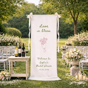 May include: A white banner with the text "Love in bloom" and "Welcome to Sophia's Bridal Shower 23.05.2026" in green and pink. The banner is decorated with a floral bouquet illustration. The scene includes wine bottles, glasses, and floral arrangements, suggesting a wedding or bridal event.