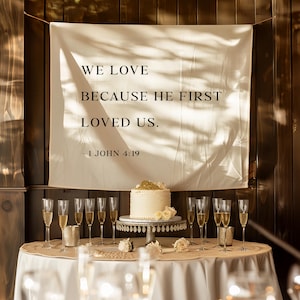 May include: A wedding reception table with a white tablecloth, champagne flutes, and a cake. A banner with the text "WE LOVE BECAUSE HE FIRST LOVED US. - 1 JOHN 4:19" hangs in the background. The cake is decorated with gold and white flowers.