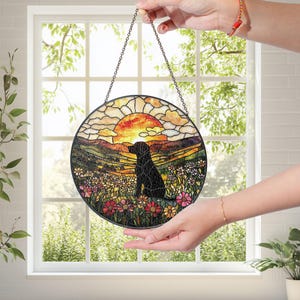 May include: Round stained glass window art featuring a black dog silhouette in a field of flowers with a sunset backdrop. The artwork is framed with a black metal border and suspended by a chain.