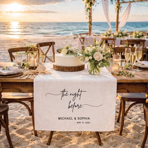 May include: A beach wedding reception table setting with a white cake on a wooden base, floral arrangements, and champagne glasses. A white table runner displays the text "the night before", along with names and a date. The scene is set on a sandy beach at sunset.