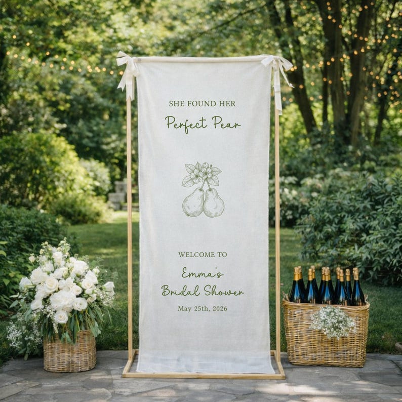 Puede incluir: Una pancarta crema con el texto "She Found Her Perfect Pear" y "Welcome to Emma's Bridal Shower, May 25th, 2026." La pancarta presenta una ilustraci&oacute;n de pera. La escena incluye un arreglo floral y una cesta de botellas de champ&aacute;n.