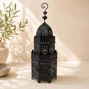 May include: A tall, black metal lantern featuring a multi-tiered, hexagonal structure with intricate, laser-cut floral and geometric patterns. The lantern is topped with a decorative finial and a curved hanging loop, creating a distinct silhouette.