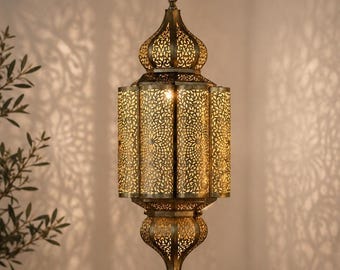 May include: A hanging pendant lamp with an antique brass finish, featuring an intricate laser-cut metal pattern that creates detailed geometric shadows. The lantern has a central cylindrical body with domed top and bottom sections, suspended by a chain.