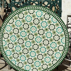 Pode incluir: Uma mesa redonda de mosaico com um padrão geométrico verde e branco. A borda da mesa é uma borda verde escura. O design do mosaico apresenta formas de estrela turquesa, verde escuro e branco. A mesa é suportada por uma base de metal escuro.