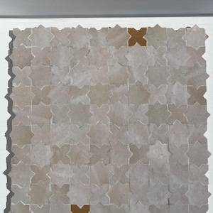 Op de afbeelding: Een vierkant van stervormige tegels in tinten beige en grijs. Twee tegels hebben een contrasterende bruine kleur. De tegels zijn gerangschikt in een rasterpatroon, waardoor een textuuroppervlak ontstaat. De tegels zijn waarschijnlijk voor vloeren of wanddecoratie.