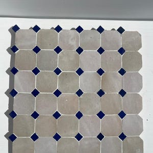 Op de afbeelding: Een vierkant tegelmozaïek met een herhalend patroon van achthoekige beige tegels met donkerblauwe vierkante accenten. De tegels zijn in een raster gerangschikt, waardoor een visueel aantrekkelijk geometrisch ontwerp ontstaat. Geschikt voor vloer- of wanddecoratie.