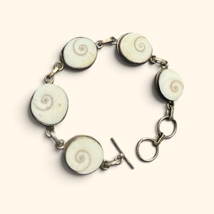 Op de afbeelding: Een zilverkleurige schakelarmband met vijf ronde, gebroken witte Shiva-oog schelp cabochons. Elke schelp vertoont een natuurlijk bruin spiraalpatroon. De armband wordt vastgemaakt met een metalen knevelsluiting bestaande uit een staafje en ring.