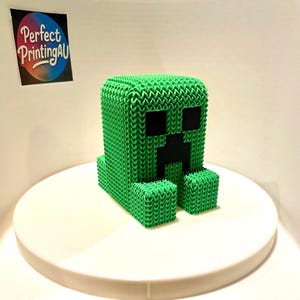 May include: A green, blocky figure with a pixelated face, resembling a popular video game character. The figure has a textured, woven appearance and is displayed on a white surface. The figure has black eyes and mouth.